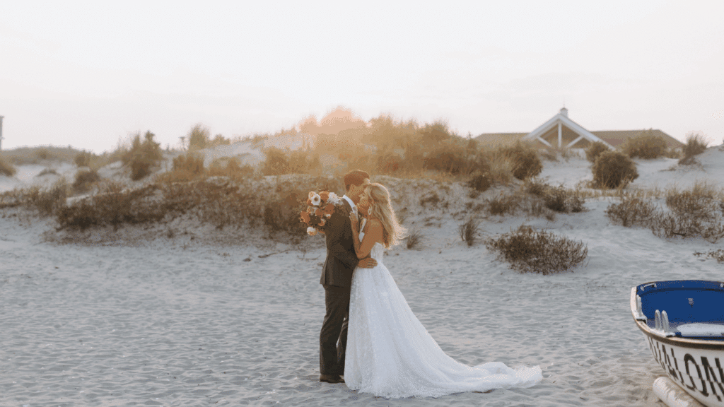 Bride and groom kissing while the sun sets behind a sand dune behind them