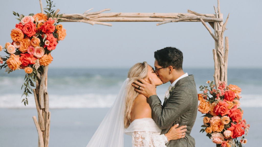 Bride and groom kissing at their beach wedding ceremony under an arch with the ocean in the background