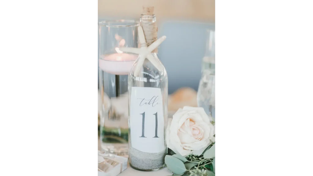Nautical themed table decor. Glass bottle with sand, sea star, and table number next to a light pink rose and floating candle.