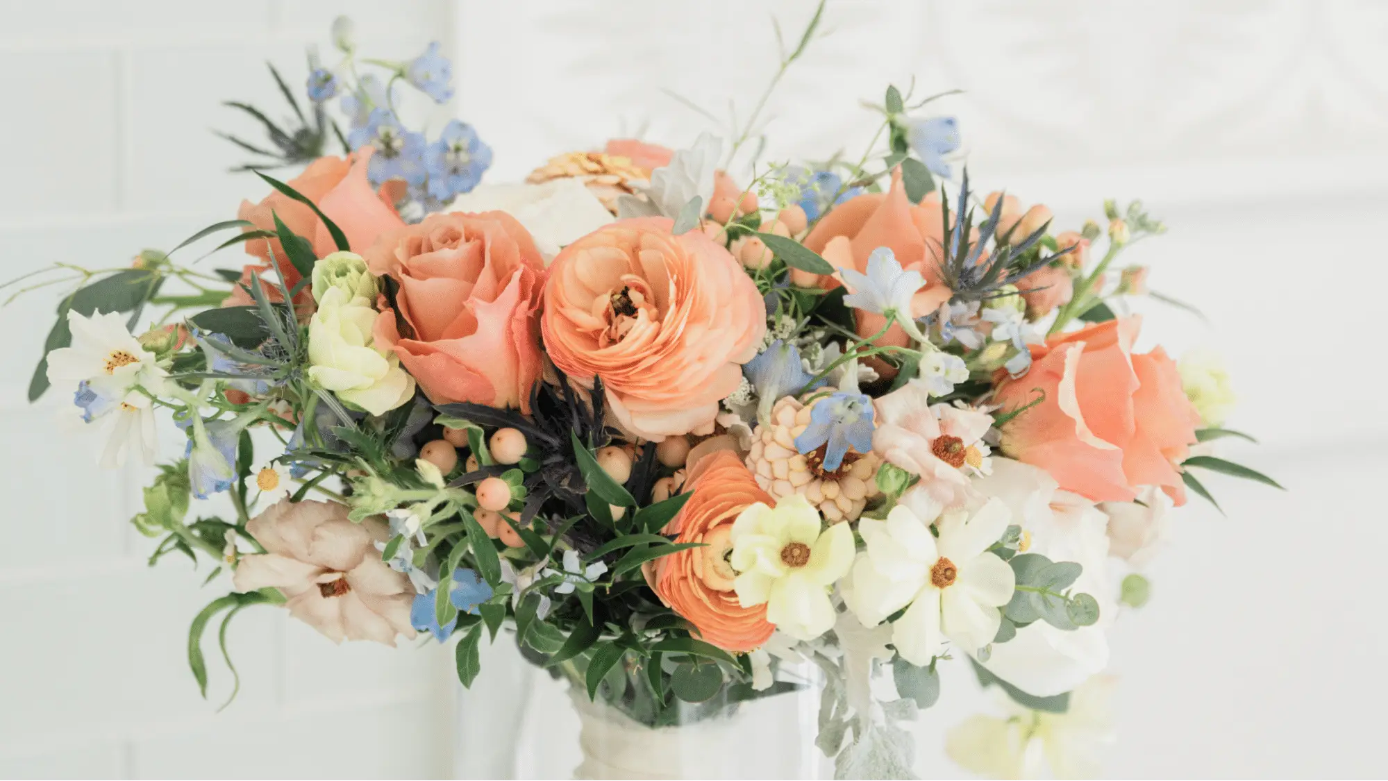 Bridal bouquet featuring peach-pink ranunculus, roses, and accents of blue and green